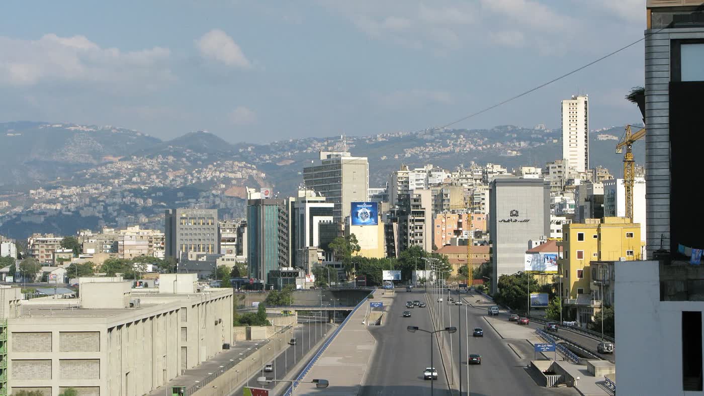 A wide Beirut road running between apartment blocks and offices, with a hazy range of hills behind