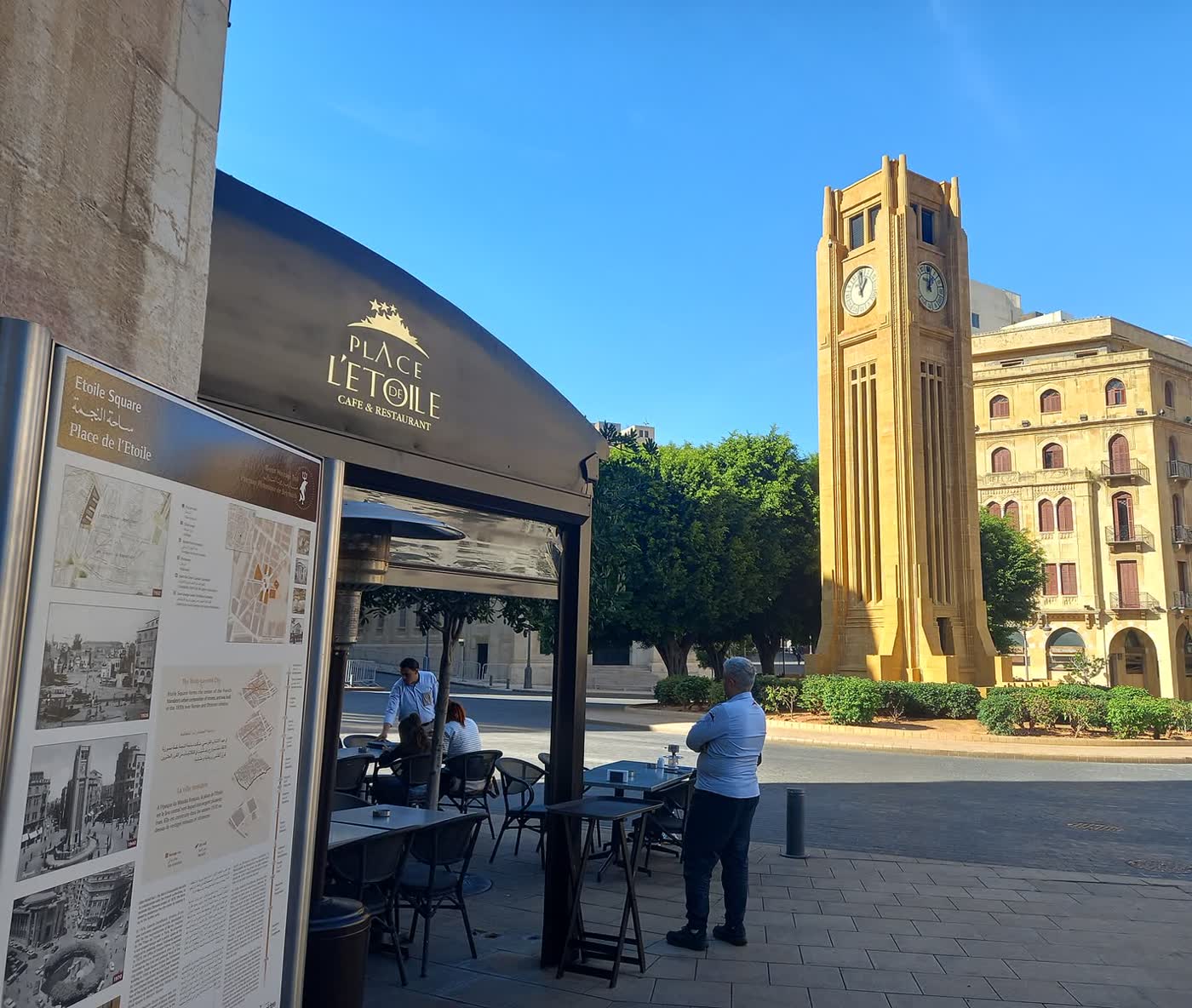 A tall sandstone clock tower on a traffic island in a sunlit square, with a café terrace and an arcaded building around it