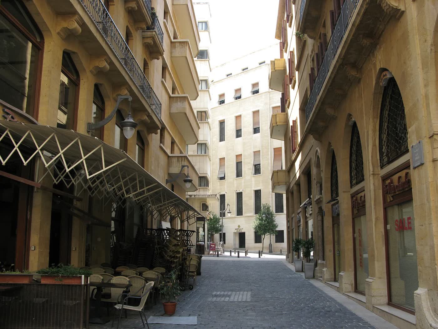A narrow cobbled street in downtown Beirut between honey-coloured stone buildings, with café chairs stacked under an awning on the left