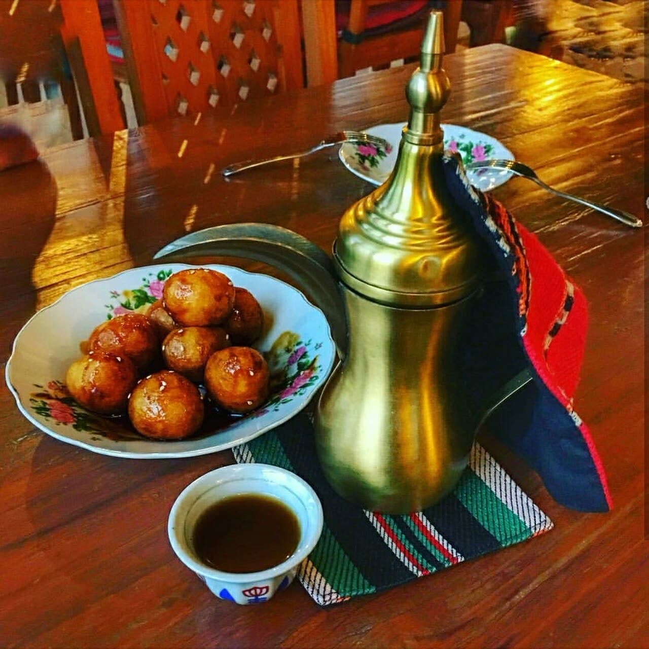 Glossy golden luqaimat in sesame seeds and syrup on a flowered plate beside a brass coffee pot and a small cup of coffee