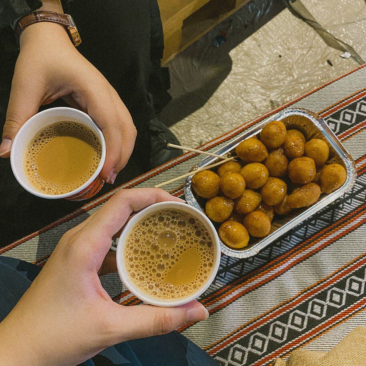 Two hands holding paper cups of karak tea above a foil tray of golden luqaimat on a woven striped mat