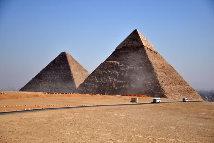 The pyramids of Giza rising from desert sand under a clear blue sky, with a road and two white vans in front