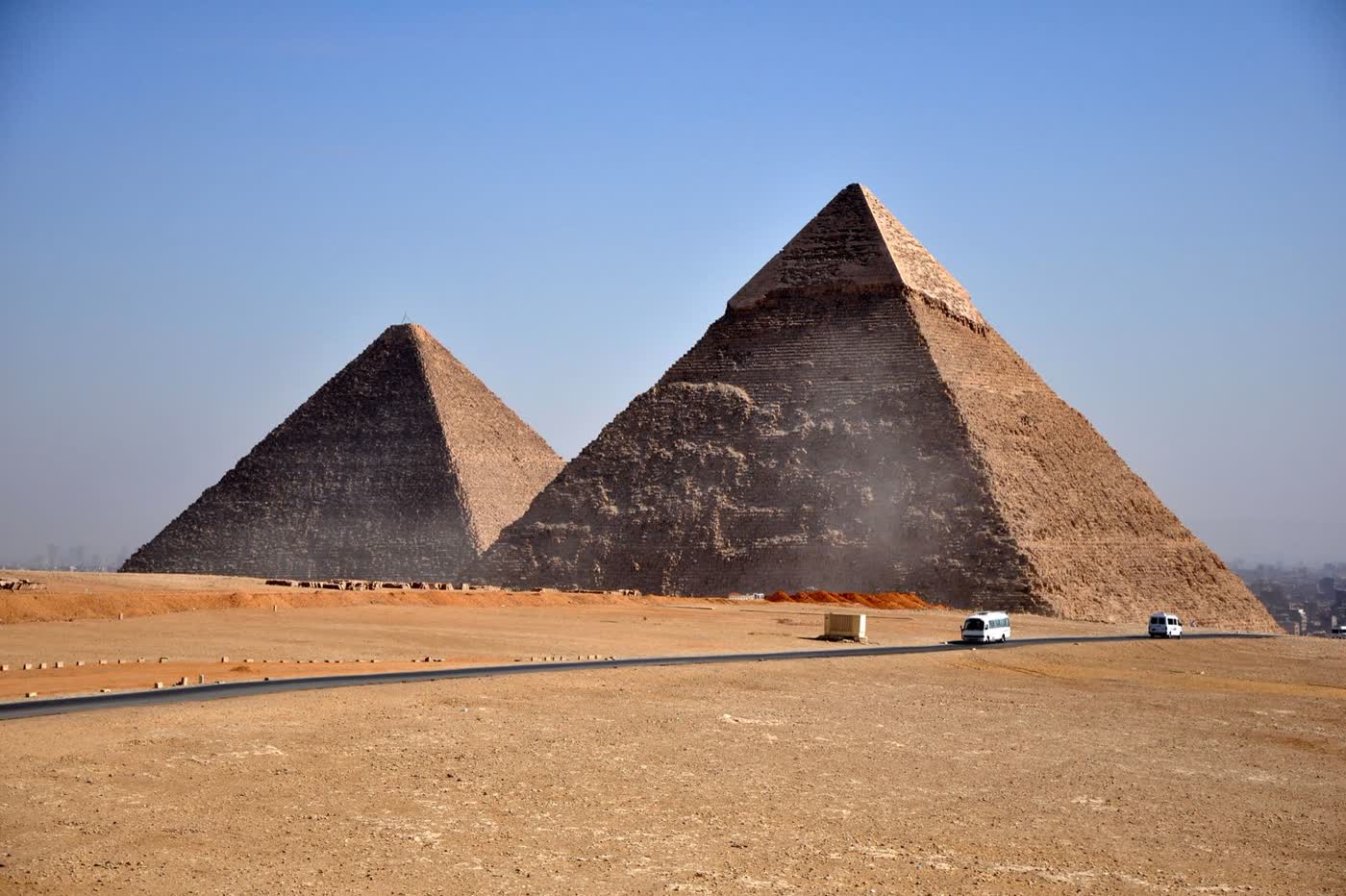 The pyramids of Giza rising from desert sand under a clear blue sky, with a road and two white vans in front