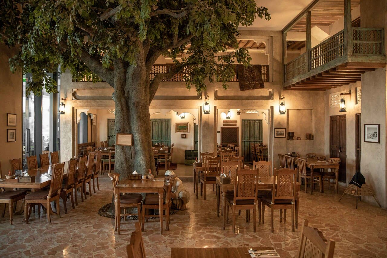A wide dining room with wooden tables and chairs around a large tree, lanterns on the walls and carved timber balconies above