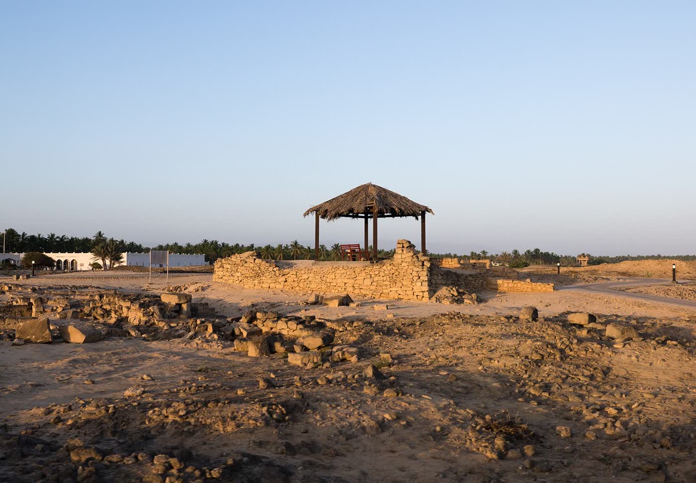 Low stone walls and a thatched shelter among ruins on flat, sandy ground, with palm trees and a white building behind them