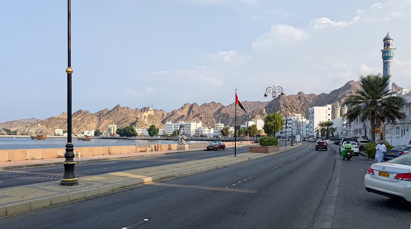 A waterfront road along a bay in Muscat, with low white buildings, a blue-tiled minaret and brown mountains behind