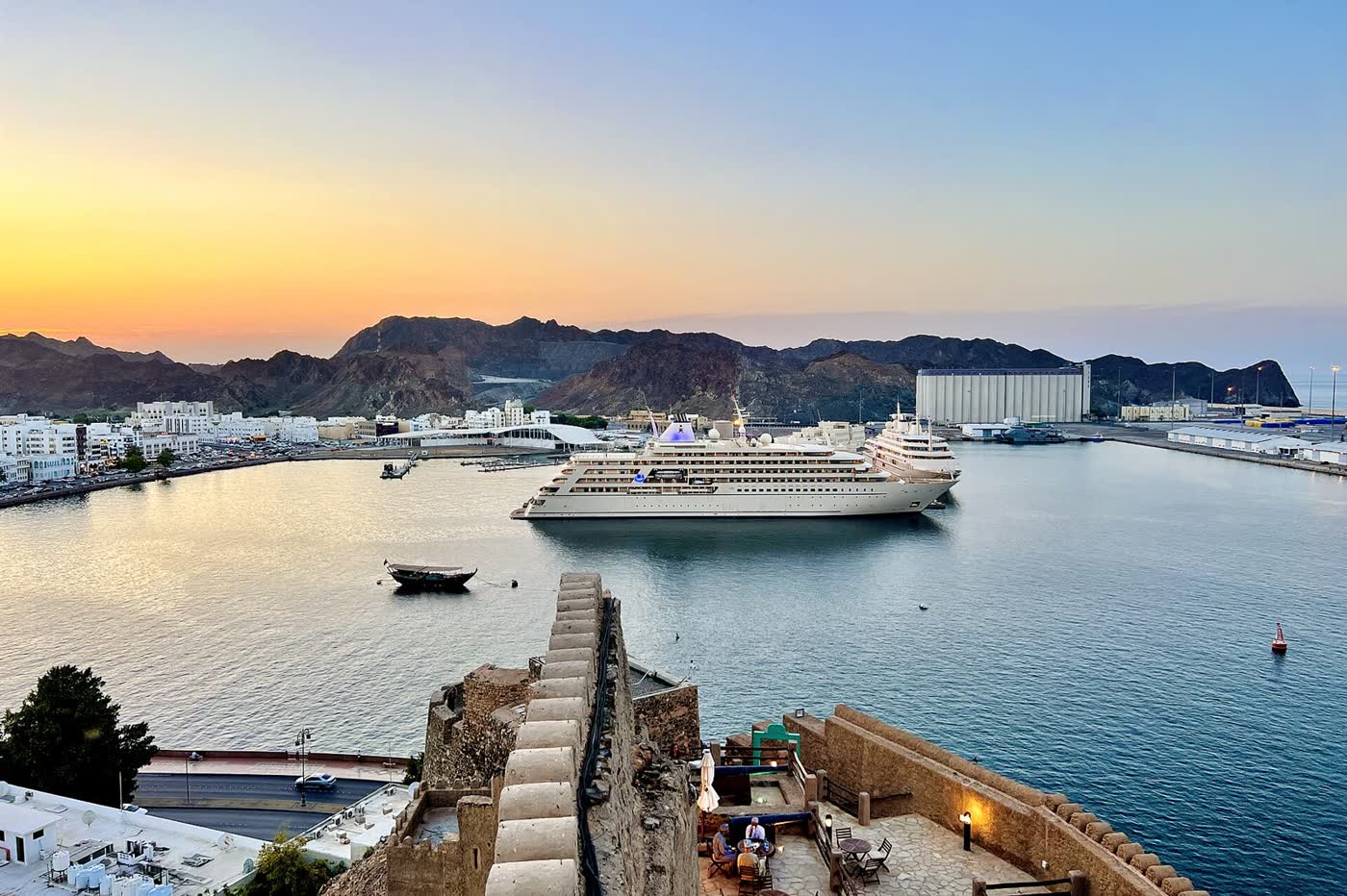 Muscat’s Sultan Qaboos Port at sunset seen from a fort wall, with a large white yacht at the quay, dhows on the water and brown mountains behind
