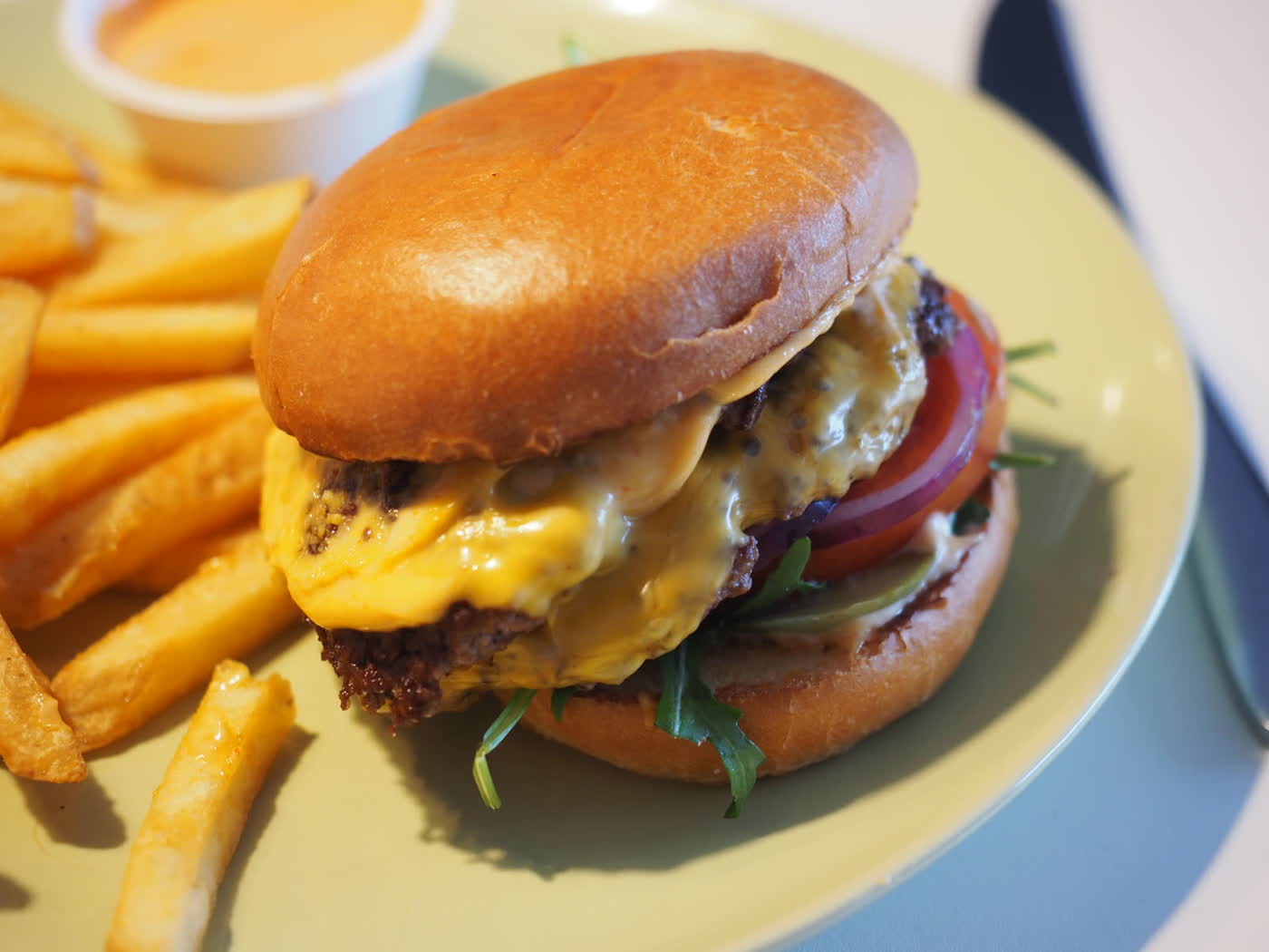 A double cheeseburger with tomato, red onion and rocket on a pale green plate beside a pile of fries