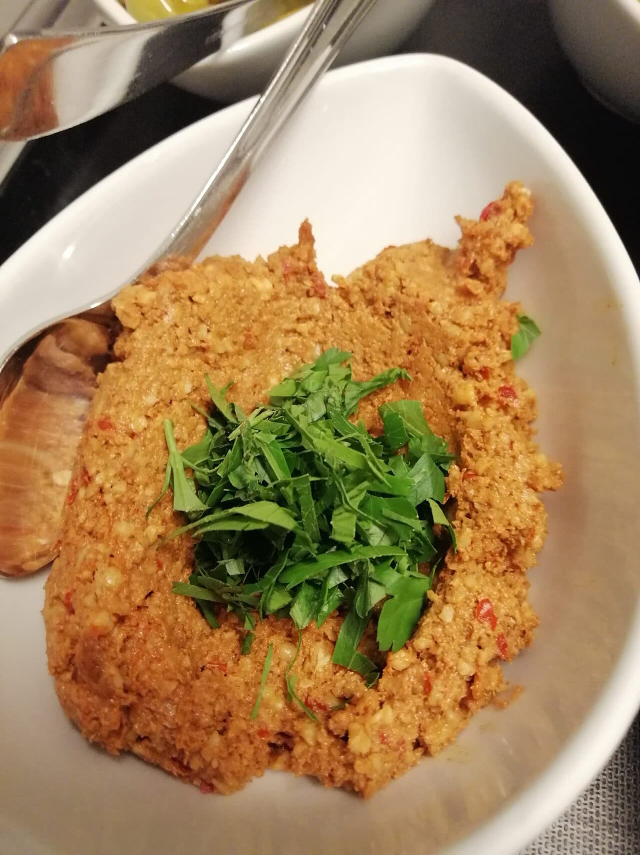 A white bowl of orange-brown walnut and red-pepper paste topped with chopped parsley, with a spoon at the side