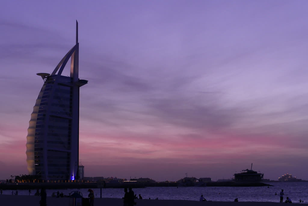 The Burj Al Arab silhouetted against a purple and pink dusk sky, seen from a public beach with people on the sand