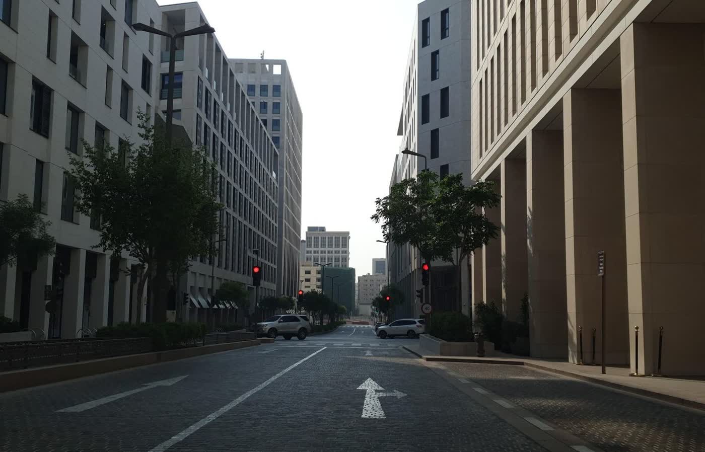 A wide paved street between pale stone office and residential buildings, with trees and a few parked cars
