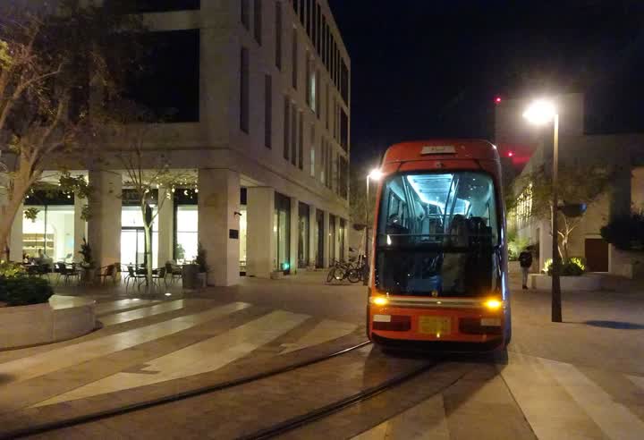 An orange tram stopped on its tracks in a stone-paved square at night, beside a pale stone building with a lit café at ground level