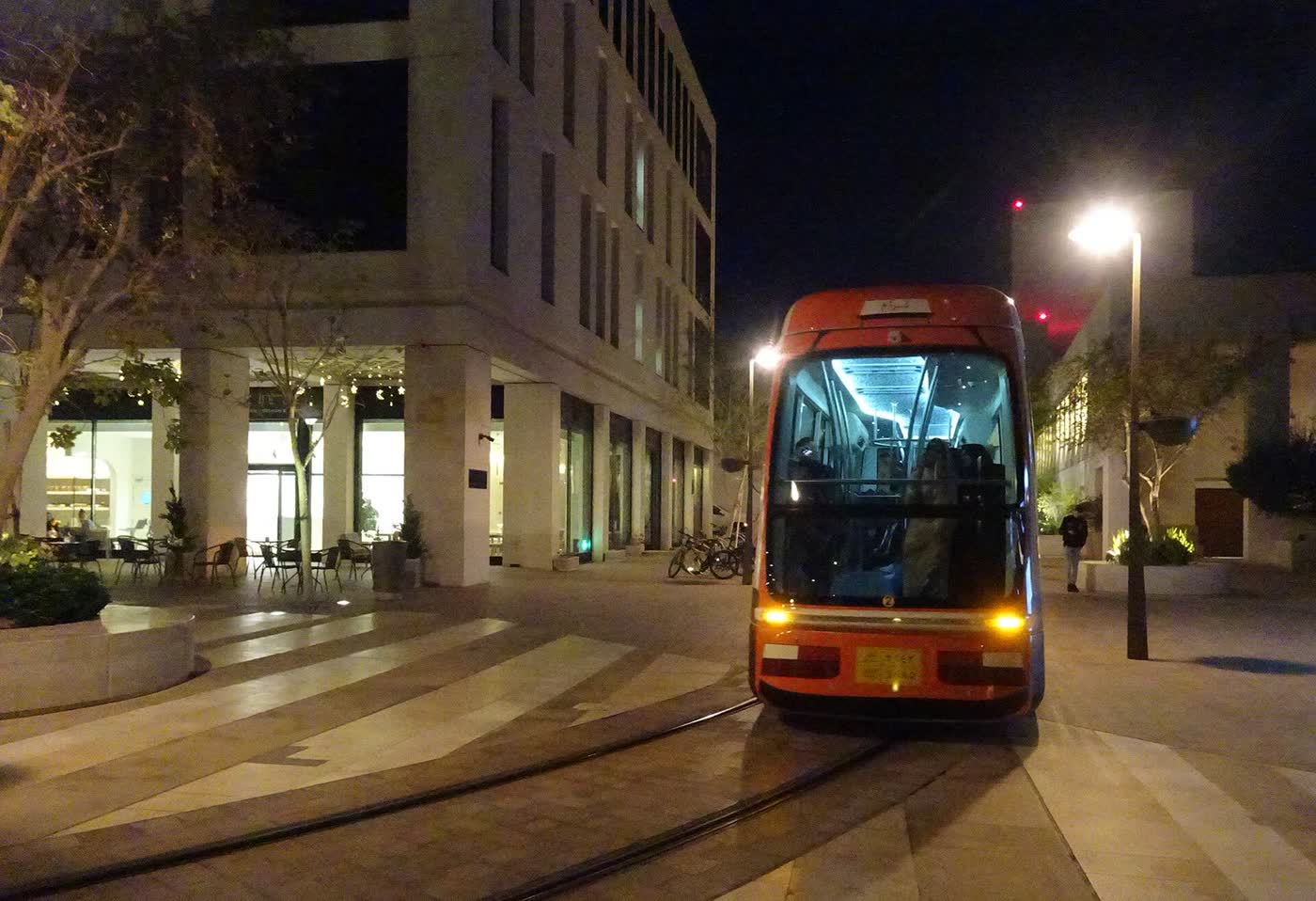 An orange tram stopped on its tracks in a stone-paved square at night, beside a pale stone building with a lit café at ground level