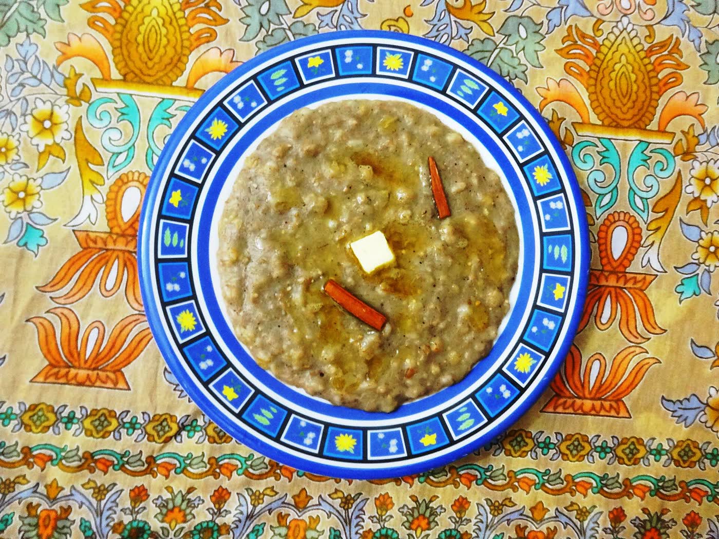 A bowl of harees, a porridge of boiled wheat, with a knob of butter and two cinnamon sticks, on a patterned tablecloth