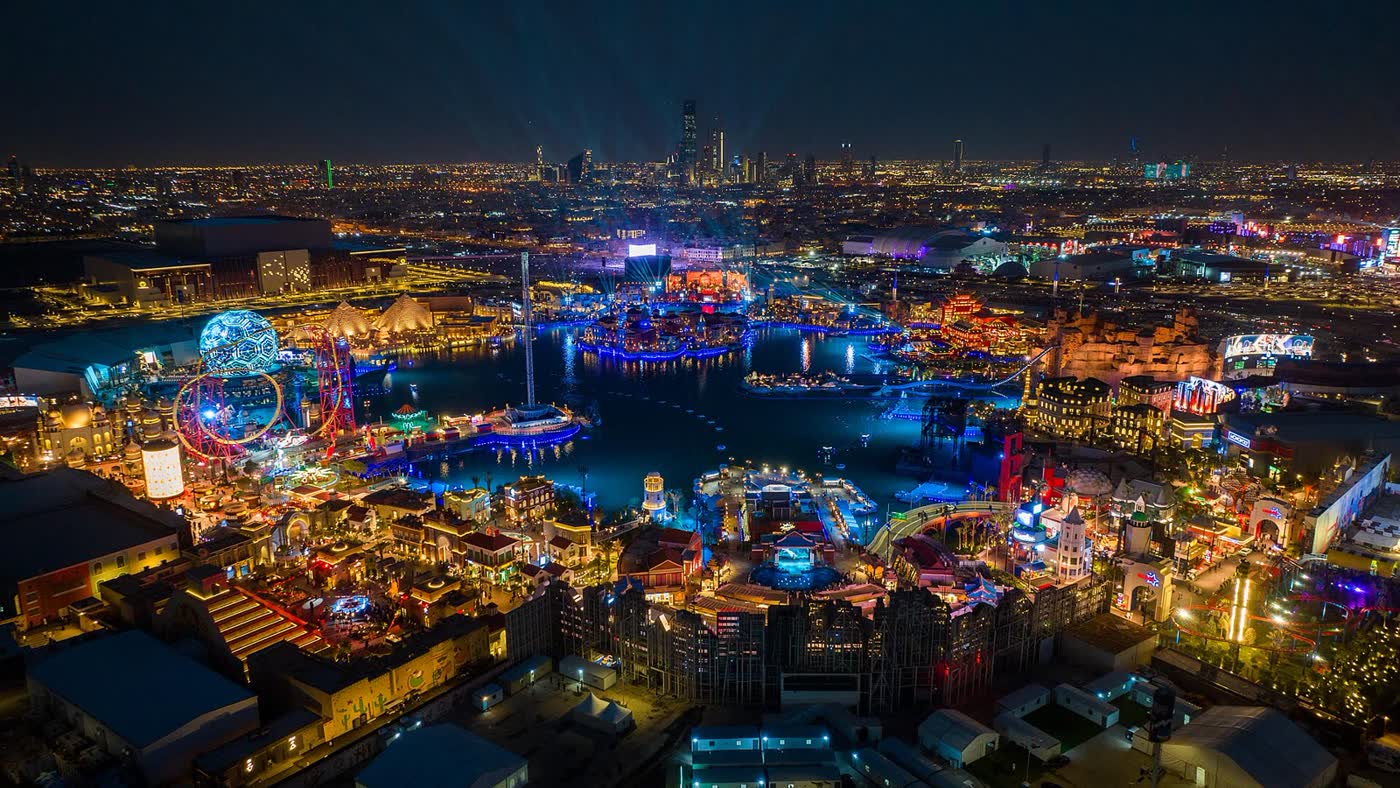Aerial view of Boulevard World in Riyadh at night, with a lake, themed pavilions, a Ferris wheel and the city skyline beyond