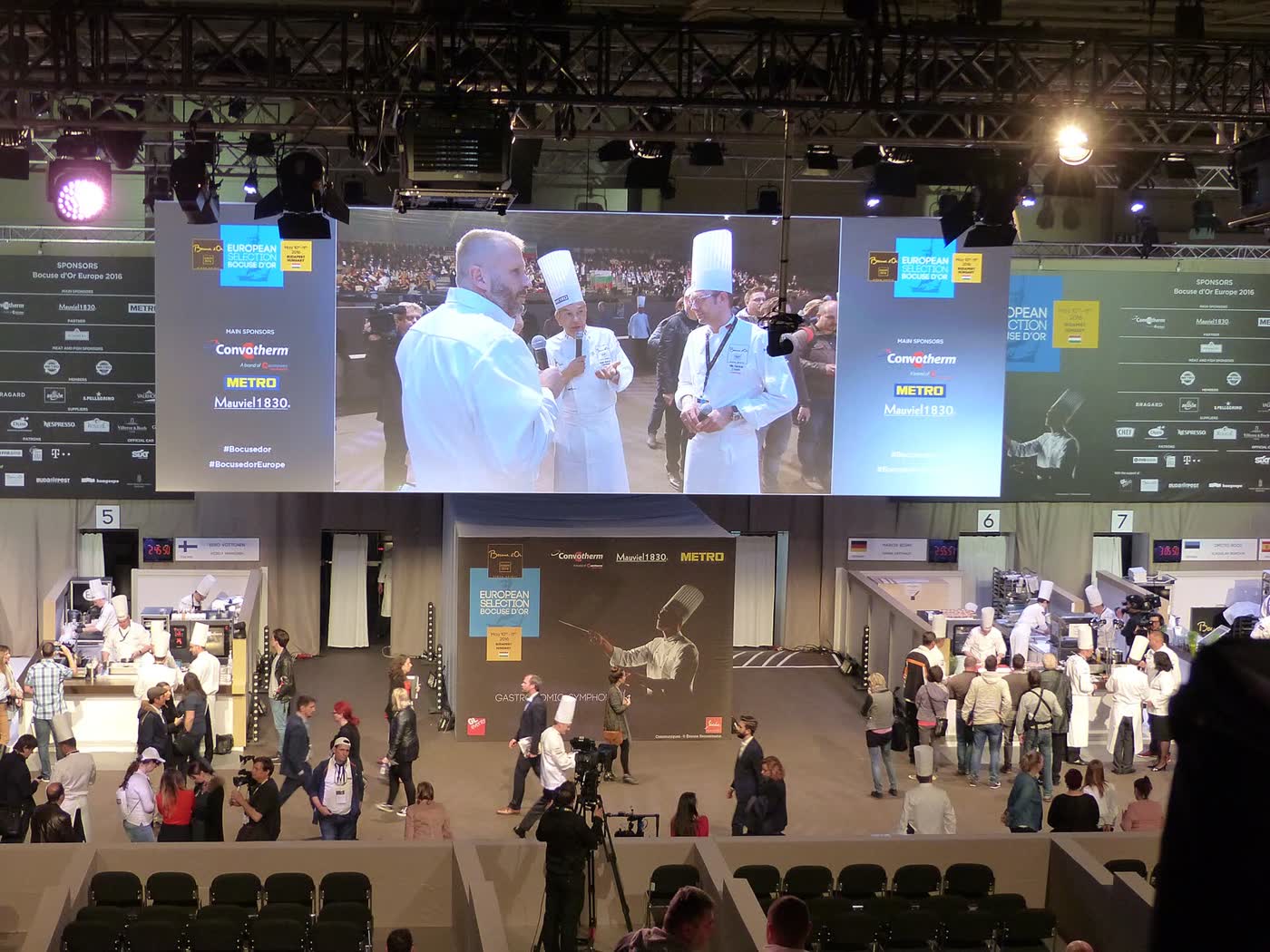 Chefs in white jackets cooking in open competition kitchens beneath a large video screen, with spectators standing below