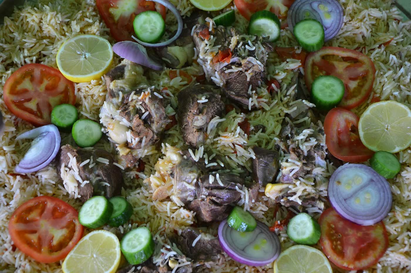 A tray of lamb kabsa seen from above, with slices of tomato, cucumber, red onion and lime scattered over the rice