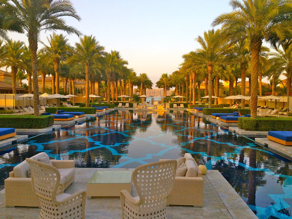 Palm trees lining a long pool with blue-and-gold tiled patterns and sofas in the foreground, at sunset